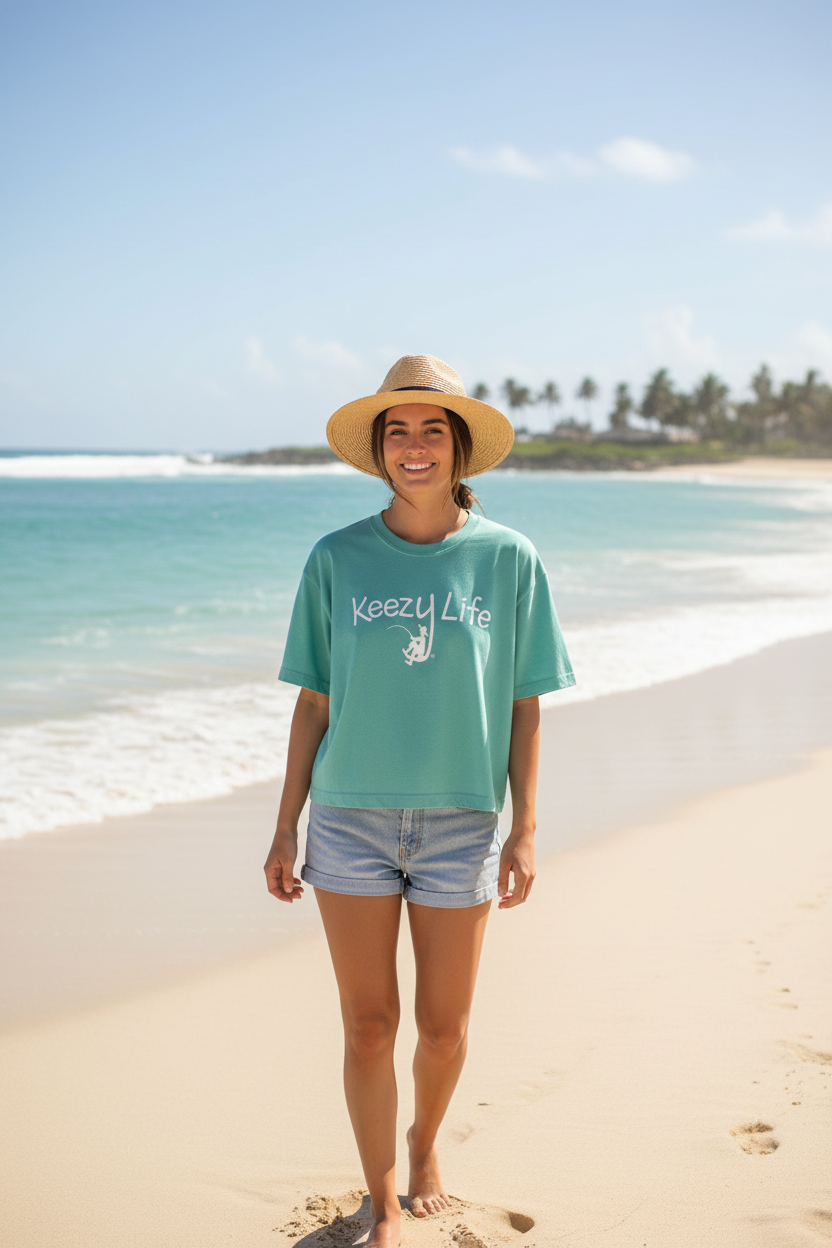 A woman stands on a sandy beach wearing a mint green graphic t-shirt with 'keezy life' printed on the front, paired with denim shorts and a straw hat.